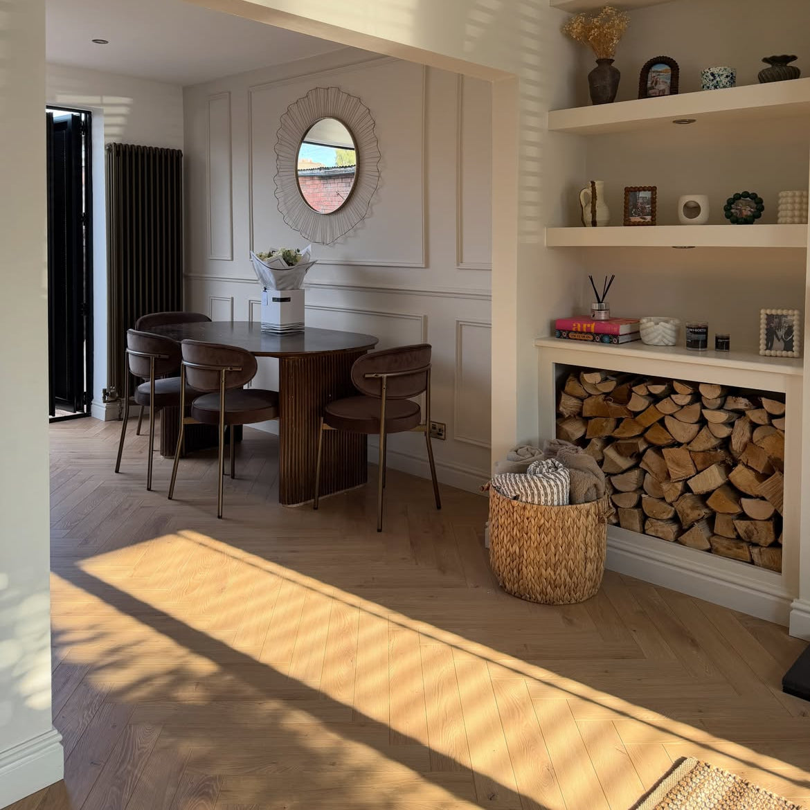 A modern dining room corner with a dark wood dining table and chairs, a round decorative mirror, built-in white shelves with decor and a firewood storage compartment, and light wood herringbone flooring with strong sunlight shadows.