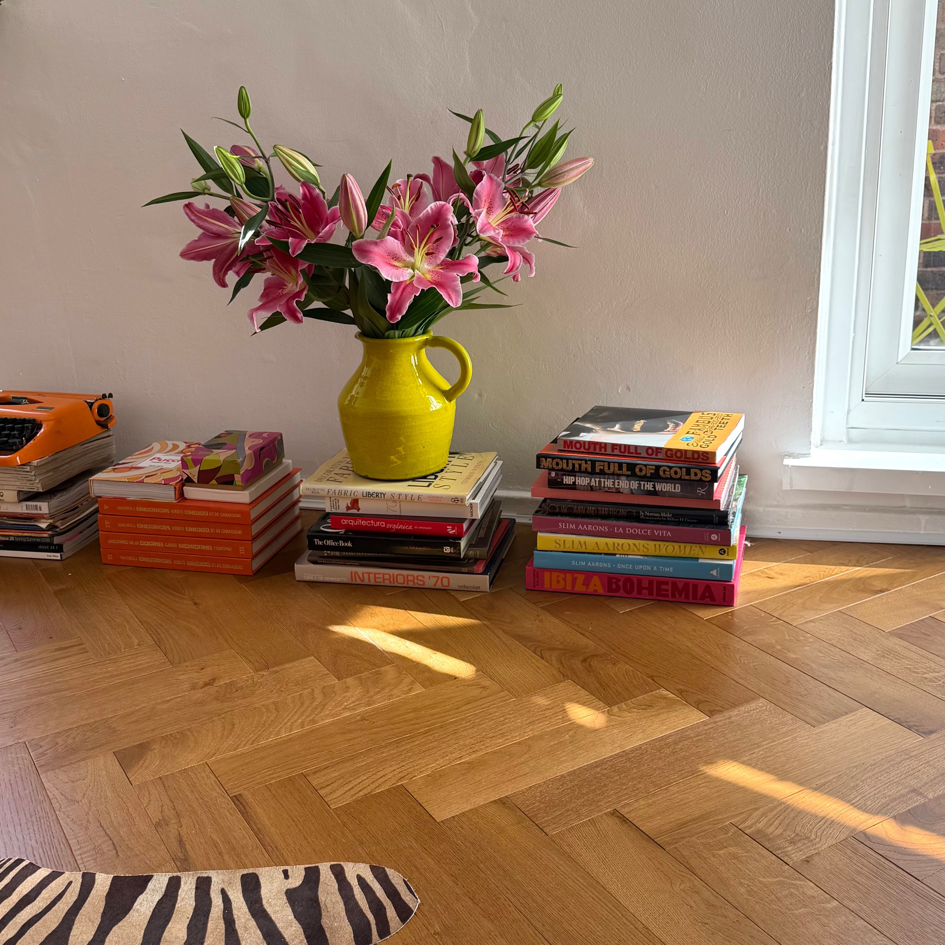 A bright yellow ceramic pitcher filled with pink lilies sits on a stack of colorful books on a light wood herringbone floor, with more books and a small orange typewriter against a white wall near a window.