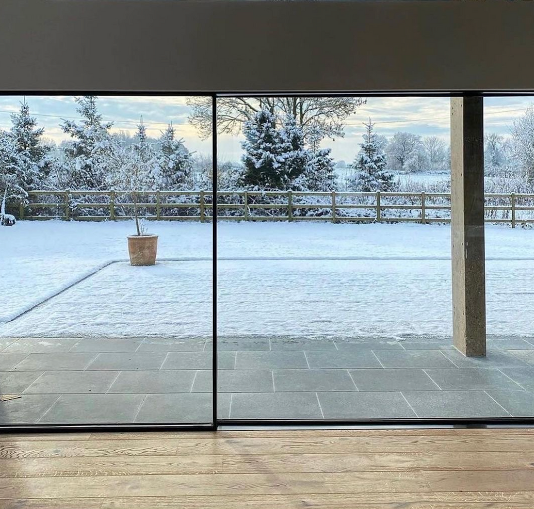 An image showcasing a warm and inviting home interior with underfloor-heated wooden flooring. The large glass windows and bifold doors at the end of the room offer a contrast with the snowy, cold outdoor scenery, accentuating the cozy indoor environment.